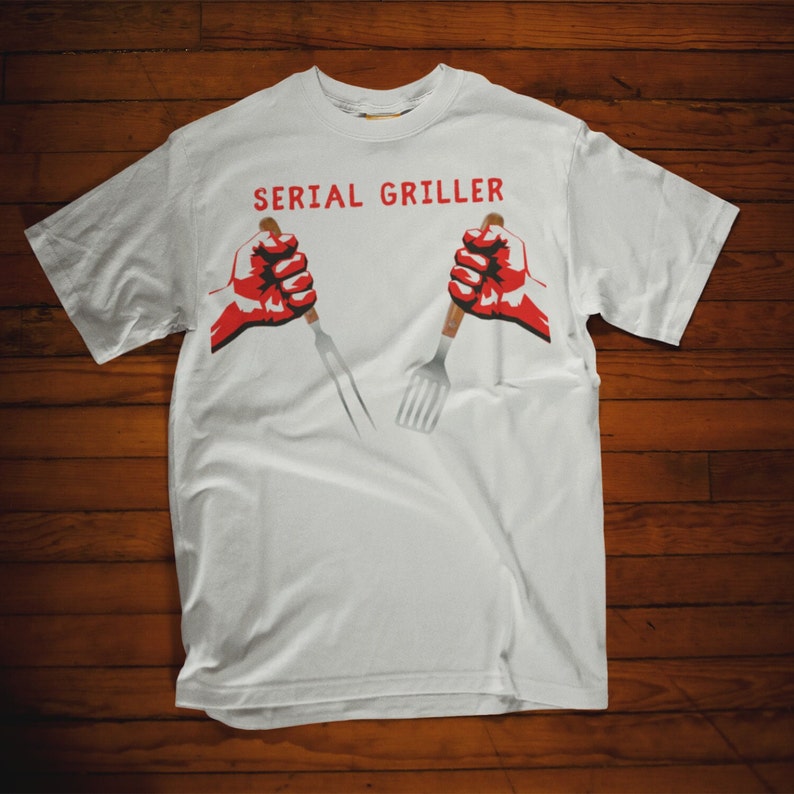 May include: White t-shirt with the text "SERIAL GRILLER" in red letters. Two red fists are holding a fork and spatula.