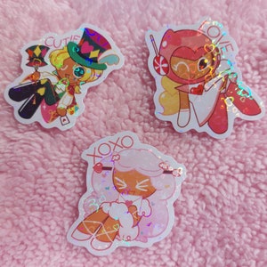 May include: Three holographic stickers featuring characters from the Cookie Run: Kingdom mobile game. The stickers are on a pink fuzzy surface. The first sticker features a character with a top hat and a playing card, the second sticker features a character with a red helmet and a lollipop, and the third sticker features a character with pink hair and a heart.