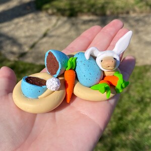 May include: A miniature Easter-themed sculpture featuring a bunny wearing a white bunny hat, blue speckled eggs, orange carrots, and a light yellow nest. One egg is broken, revealing a chocolate interior. A small white ball is also present.