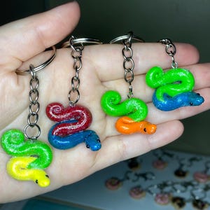 May include: Four colorful snake-shaped keychains. Each keychain features a different color combination: green and yellow, red and blue, green and orange, and green and blue. The snakes are made of a glossy material and attached to silver keyrings.
