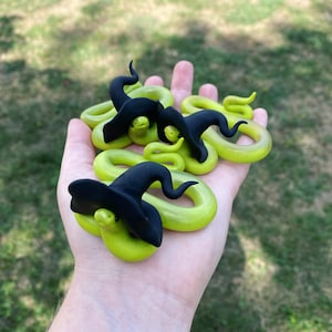 May include: A collection of small, coiled, lime green snake figurines, each wearing a black witch hat. The snakes have yellow eyes and are arranged in a hand, with a blurred green background.