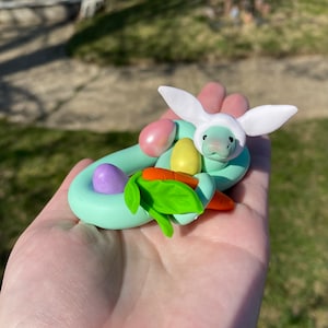 May include: A whimsical, handmade polymer clay sculpture of a snake wearing a bunny hat. The snake is light green and coiled around colorful Easter eggs and carrots. The bunny hat is white, and the eggs are pink, yellow, and purple.