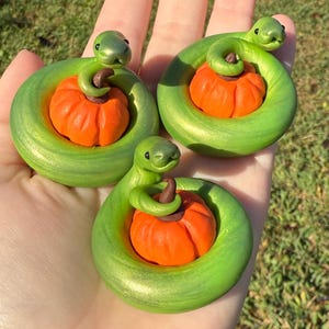 May include: Three handmade green snake figurines coiled around orange pumpkins. The snakes have detailed features and are positioned on a light-colored surface. The pumpkins have a textured surface and a brown stem. The overall theme is Halloween.