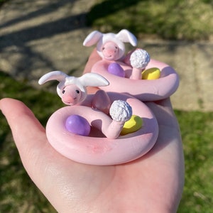 May include: Two pink clay snake figurines, each wearing a bunny hat. The snakes coil around colorful Easter eggs and white mushroom-shaped decorations. The figurines are held in a hand, with a blurred green background.