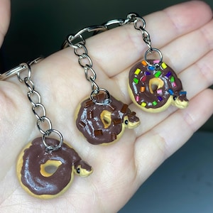 May include: Three handmade donut-shaped keychains with a chocolate glaze and sprinkles. Each keychain features a small, snake-like creature emerging from the donut. The keychains are attached to silver-colored chains and key rings.