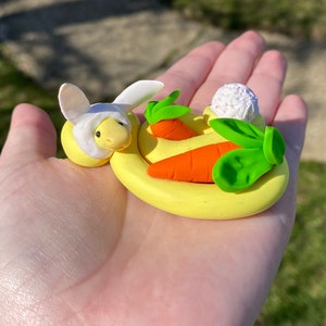 May include: A yellow clay sculpture of a turtle wearing a white bunny hat, with orange carrots and green leaves on a yellow base. A white ball of clay is also on the base. The sculpture is held in a hand.