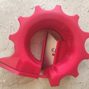 Op de afbeelding: Een rood plastic gereedschap met een tandwielvormig handvat en een metalen mes. Het gereedschap is ontworpen om tape te snijden.