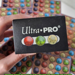 May include: A black Ultra-PRO deck box with a silver logo and three colored circles. The circles are red, green, and yellow. The text "Ultra-PRO&reg;" is printed on the box. "STICKERS SHOWN FOR SCALE ON ULTRA-PRO DECK BOX. DECK BOX NOT INCLUDED" is printed below the circles.
