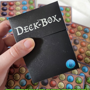 May include: A black DECK-BOX card box with a blue water drop sticker on the front. The text "STICKERS SHOWN FOR SCALE ON ULTRA-PRO DECK BOX. DECK BOX NOT INCLUDED" is printed below the box.