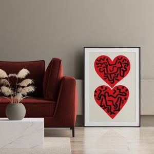 以下が含まれることがあります： 黒い抽象的なデザインが描かれた赤いハートが2つ入った額装されたプリント。ハートはクリーム色の背景に描かれています。プリントは赤いソファの上の壁に掛けられています。