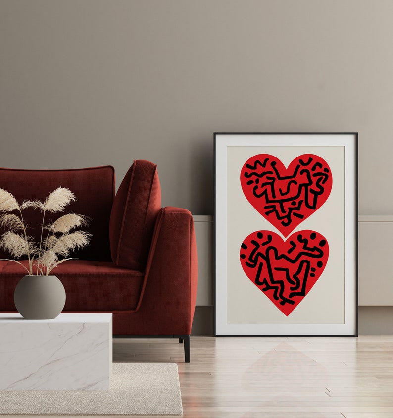 以下が含まれることがあります： 黒い抽象的なデザインが描かれた赤いハートが2つ入った額装されたプリント。ハートはクリーム色の背景に描かれています。プリントは赤いソファの上の壁に掛けられています。