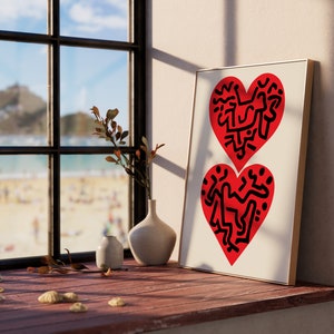以下が含まれることがあります： 白い背景に黒い抽象的なデザインが施された赤いハート2つが描かれた額装されたプリント。プリントは、花瓶と小さな植物のある木の窓枠の上に置かれています。