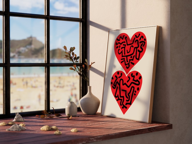 以下が含まれることがあります： 白い背景に黒い抽象的なデザインが施された赤いハート2つが描かれた額装されたプリント。プリントは、花瓶と小さな植物のある木の窓枠の上に置かれています。