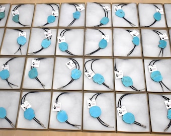May include: A collection of thirty-two bolo ties, each featuring a large, irregular, bright turquoise stone centerpiece on a black braided cord with silver-toned metal tips, individually packaged in square boxes with white foam inserts.