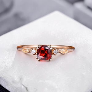 May include: A rose gold ring with a square, deep red gemstone at its centre, with two smaller white gemstones either side. The band has decorative accents. A delicate piece of jewellery.