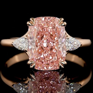 May include: A rose gold ring featuring a large, rectangular pink diamond as the centerpiece, flanked by two pear-shaped white diamonds. The ring's setting has intricate detailing, and the background is black.