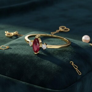 May include: A gold ring with a marquise-cut ruby center stone and two smaller diamond accents. The ring is set on a dark green velvet surface, with gold jewelry and a pearl in the background. The ring is a piece of fine jewelry.