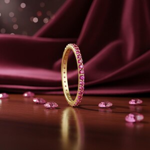 May include: A gold ring featuring a continuous line of small, bright pink gemstones. The ring is displayed against a backdrop of deep burgundy fabric and scattered pink gemstones, creating a luxurious and elegant aesthetic.