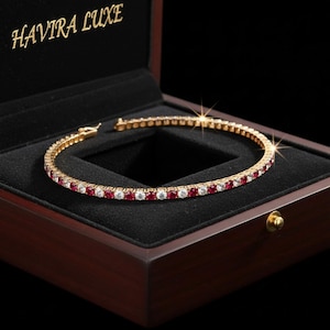 Puede incluir: Una pulsera de tenis dorada con piedras preciosas rojas y blancas alternas, exhibida en un joyero de madera oscura. El joyero está forrado con terciopelo negro y tiene las palabras "HAVIRA LUXE" en letras doradas en el interior de la tapa.