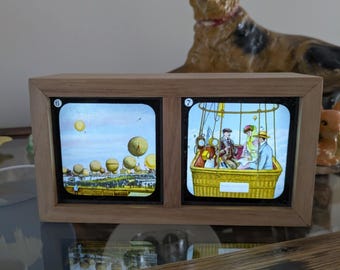 Könnte beinhalten: Ein Vintage-Stereoskop mit Holzrahmen und zwei beleuchteten Bildern. Ein Bild zeigt eine Gruppe von Heißluftballons, das andere Menschen in einem Heißluftballonkorb. Der Rahmen besteht aus hellfarbigem Holz.