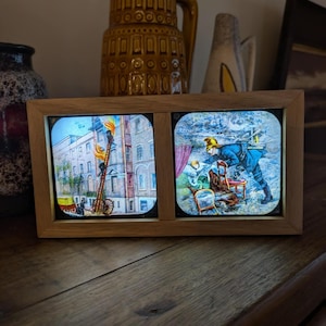May include: A wooden framed diptych with two illuminated slides. One shows a street scene with a fire ladder, the other a firefighter assisting someone. The frame rests on a wooden surface, with decorative vases in the background.