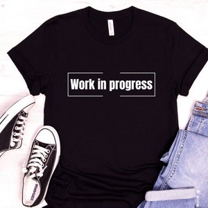 Puede incluir: Camiseta negra con el texto "Work in progress" impreso en blanco en la parte delantera. La camiseta está colocada sobre una superficie blanca con un par de zapatillas negras y unos vaqueros azules.