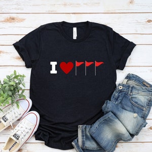 Puede incluir: Una camiseta azul marino con un gráfico blanco "I love" y tres banderas rojas. La camiseta está sobre un fondo de madera blanca con un par de zapatillas blancas y unos vaqueros azules.