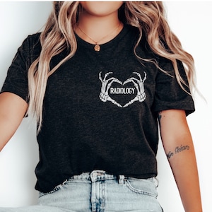 Op de afbeelding: Een zwart T-shirt met een afbeelding van twee skelethandjes die een hart vormen met het woord "RADIOLOGY" in het hart.