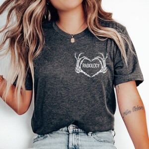 Op de afbeelding: Een donkergrijs T-shirt met een witte afbeelding van twee skelethandjes die een hart vormen met het woord "RADIOLOGY" erin.