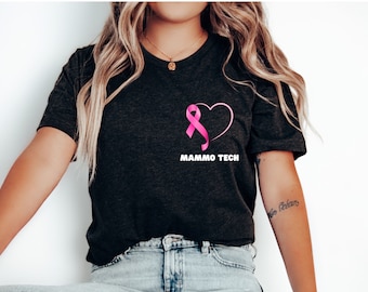 Peut inclure: T-shirt gris foncé avec un ruban rose à l'intérieur d'un cœur et le texte blanc « MAMMO TECH ». Le t-shirt a un col en V et des manches courtes. Le motif est sur la poitrine gauche.