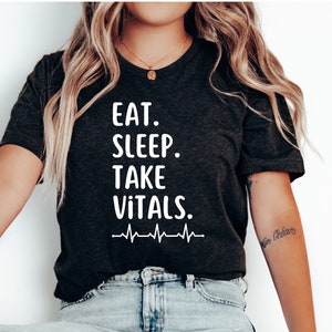 Puede incluir: Una camiseta gris oscuro con el texto "EAT. SLEEP. TAKE VITALS." en letras blancas y un gráfico de latido del corazón blanco debajo del texto.