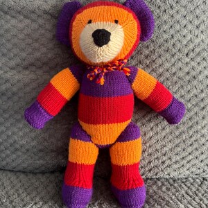 May include: A handmade knitted bear in vibrant colors. The bear features a cream-colored face, black nose, and a colorful scarf. The body is striped with orange, red, and purple. The bear's arms, legs, and ears are also in matching colors.