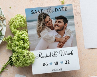Könnte beinhalten: Save-the-Date-Karte mit einem Foto eines Paares am Strand. Die Karte ist weiß mit blauen Akzenten und trägt den Text "Save the Date" in schwarzer Schrift. Die Namen des Paares sind Rachel & Mark, und das Datum ist der 06. 18. 22. Der Ort ist Atlanta, GA.