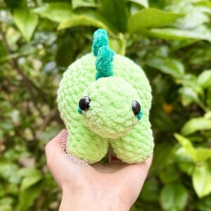 May include: A light green crocheted dinosaur plush toy with a textured yarn finish, dark round plastic eyes, and a darker green spiky ridge along its back. The toy features the text AQCROCHETS printed in a small, light-colored font on its side.