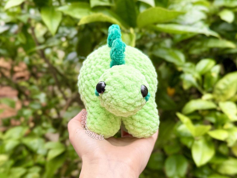 May include: A light green crocheted dinosaur plush toy with a textured yarn finish, dark round plastic eyes, and a darker green spiky ridge along its back. The toy features the text AQCROCHETS printed in a small, light-colored font on its side.