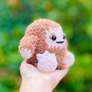 Op de afbeelding: Een handgemaakt, gehaakt pluche wezen in bruin- en wittinten, met kleine zwarte ogen en een gestikte mond. Het zachte, getextureerde speelgoed heeft kleine armen en voeten en wordt in een hand gehouden tegen een vage groene achtergrond. Het speelgoed is een grillig, knuffelbaar item.