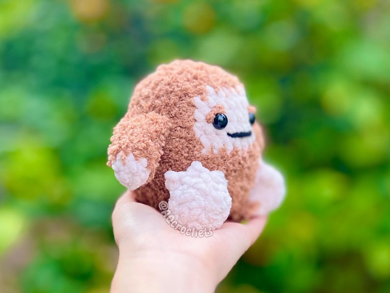 Op de afbeelding: Een handgemaakt, gehaakt pluche wezen in bruin- en wittinten, met kleine zwarte ogen en een gestikte mond. Het zachte, getextureerde speelgoed heeft kleine armen en voeten en wordt in een hand gehouden tegen een vage groene achtergrond. Het speelgoed is een grillig, knuffelbaar item.
