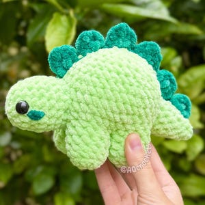 May include: A light green crochet dinosaur plush toy with a textured, soft yarn finish. The dinosaur features a row of dark green, rounded spikes along its back, a small tail, and two black plastic safety eyes with small dark green cheek patches.