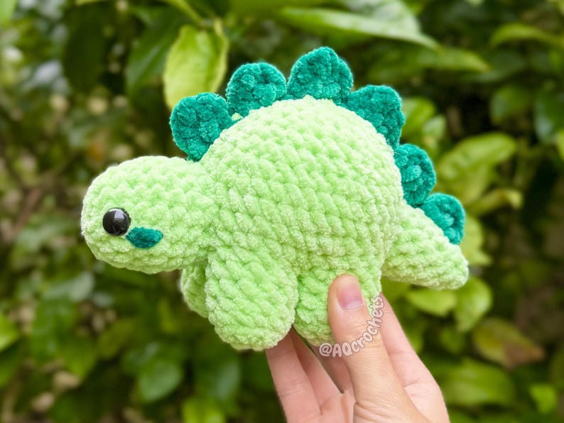 May include: A light green crochet dinosaur plush toy with a textured, soft yarn finish. The dinosaur features a row of dark green, rounded spikes along its back, a small tail, and two black plastic safety eyes with small dark green cheek patches.
