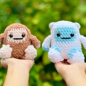 Op de afbeelding: Twee handgemaakte gehaakte pluche speelgoed. E&eacute;n is bruin met een cr&egrave;mekleurig gezicht en beige voeten en armen. De andere is wit met blauwe accenten. Beide hebben zwarte ogen en lachende monden. Het speelgoed wordt tegen een vage groene achtergrond gehouden.