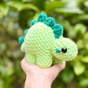 May include: A light green crocheted dinosaur plush toy with a textured yarn finish, featuring dark green dorsal plates along its back, a small tail, and black plastic safety eyes. The text @ancrochet is visible on the side of the dinosaur.