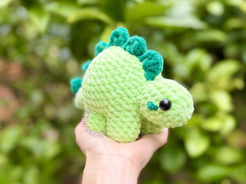 May include: A light green crocheted dinosaur plush toy with a textured yarn finish, featuring dark green dorsal plates along its back, a small tail, and black plastic safety eyes. The text @ancrochet is visible on the side of the dinosaur.