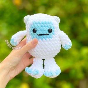 Op de afbeelding: Een handgemaakte witte en lichtblauwe gehaakte yeti-pop met zwarte veiligheidsoogjes en een gestikt lachje. De pop heeft kleine armen en benen en wordt voor een vage groene achtergrond gehouden. De pop is ongeveer 13 cm hoog.
