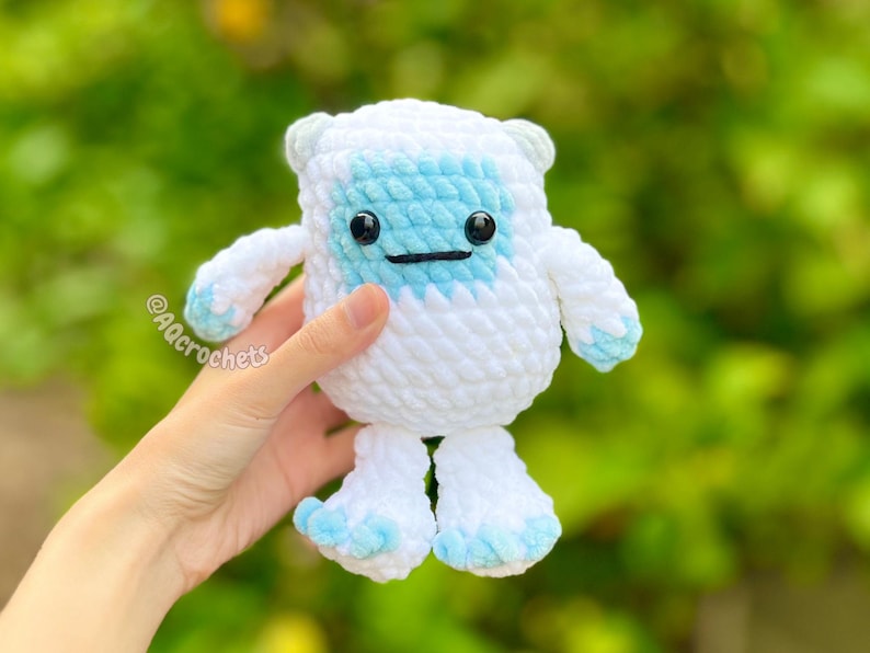 Op de afbeelding: Een handgemaakte witte en lichtblauwe gehaakte yeti-pop met zwarte veiligheidsoogjes en een gestikt lachje. De pop heeft kleine armen en benen en wordt voor een vage groene achtergrond gehouden. De pop is ongeveer 13 cm hoog.