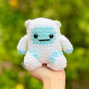 Op de afbeelding: Een handgemaakte, wit en blauw gehaakte yeti-knuffel met zwarte veiligheidsoogjes en een gestikte glimlach. De knuffel heeft een lichtblauw gezicht, voeten en accenten. Het item wordt in een hand gehouden tegen een vage groene achtergrond. De naam van de kunstenaar is zichtbaar.