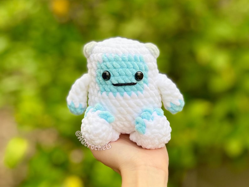 Op de afbeelding: Een handgemaakte, wit en blauw gehaakte yeti-knuffel met zwarte veiligheidsoogjes en een gestikte glimlach. De knuffel heeft een lichtblauw gezicht, voeten en accenten. Het item wordt in een hand gehouden tegen een vage groene achtergrond. De naam van de kunstenaar is zichtbaar.