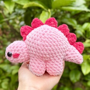 May include: A small, handmade crochet dinosaur plush toy made from soft, light pink yarn. It features a row of bright red, textured spikes along its back, a rounded head with a single black plastic safety eye, and a small embroidered red mouth.