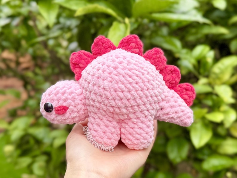 May include: A small, handmade crochet dinosaur plush toy made from soft, light pink yarn. It features a row of bright red, textured spikes along its back, a rounded head with a single black plastic safety eye, and a small embroidered red mouth.