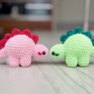 May include: Two small crocheted dinosaur plushies. One is light pink with a dark red spine, and the other is light green with a dark green spine. Both have black plastic eyes and the text @Aqcrochets printed near the pink dinosaur's feet.