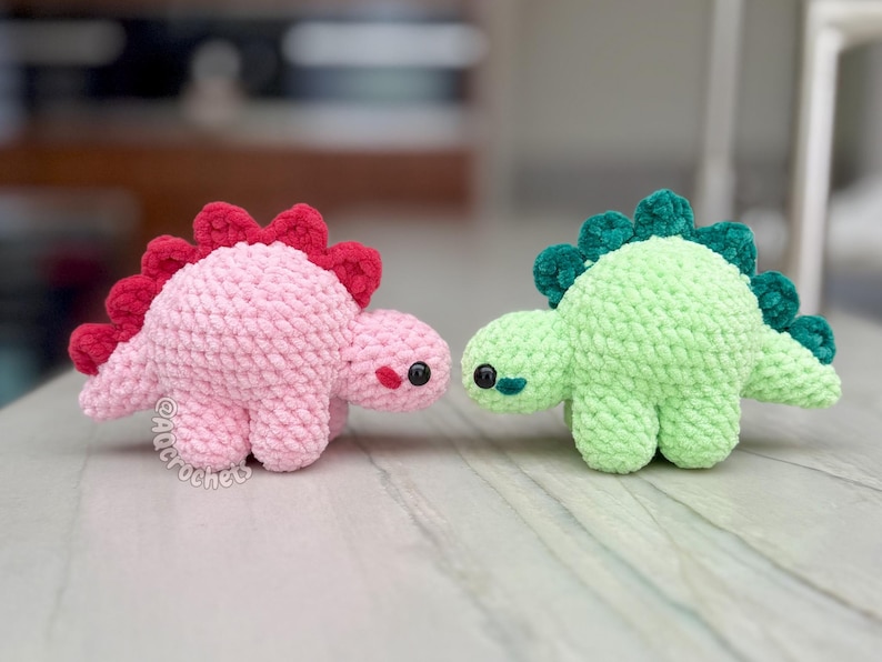 May include: Two small crocheted dinosaur plushies. One is light pink with a dark red spine, and the other is light green with a dark green spine. Both have black plastic eyes and the text @Aqcrochets printed near the pink dinosaur's feet.